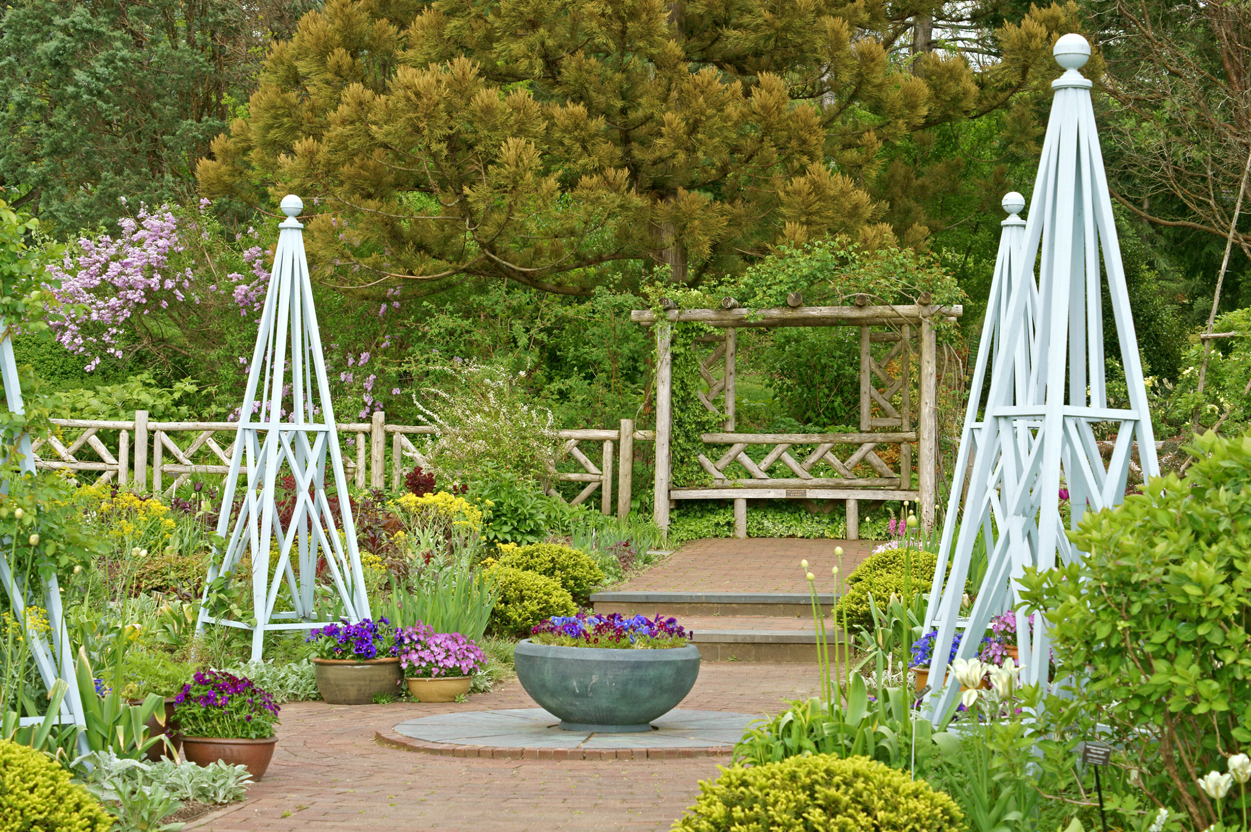 A scenic stone path at Wave Hill Public Garden featuring vibrant purple pansies, blue wooden obelisks, and a rustic wooden pergola, showcasing the lush green spaces found in the best neighborhoods in the Bronx.