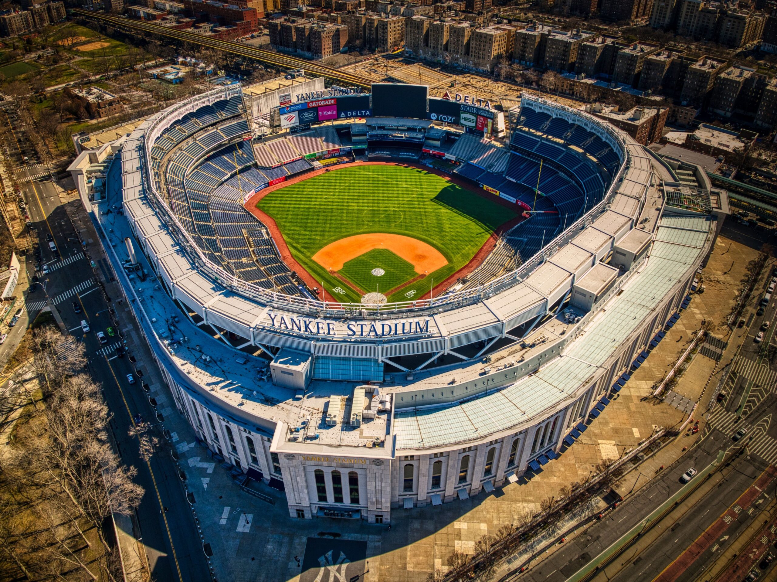 An aerial view of the iconic Yankee Stadium in late afternoon sun, showing the pristine green baseball field and surrounding city streets in one of the best neighborhoods in the Bronx.