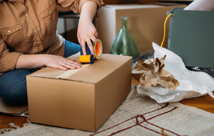 A person sits on the floor taping a cardboard box closed while packing to move.
