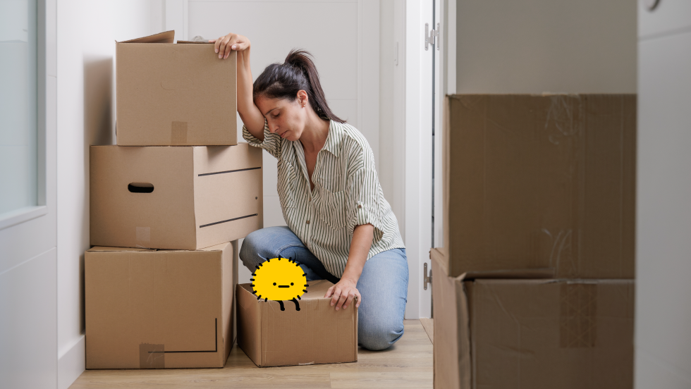 A woman looking exhausted and overwhelmed leans her head against a stack of cardboard moving boxes in a hallway, illustrating the stress of managing spring moving costs and logistics. A small, round yellow mascot character sits on top of one of the boxes next to her.